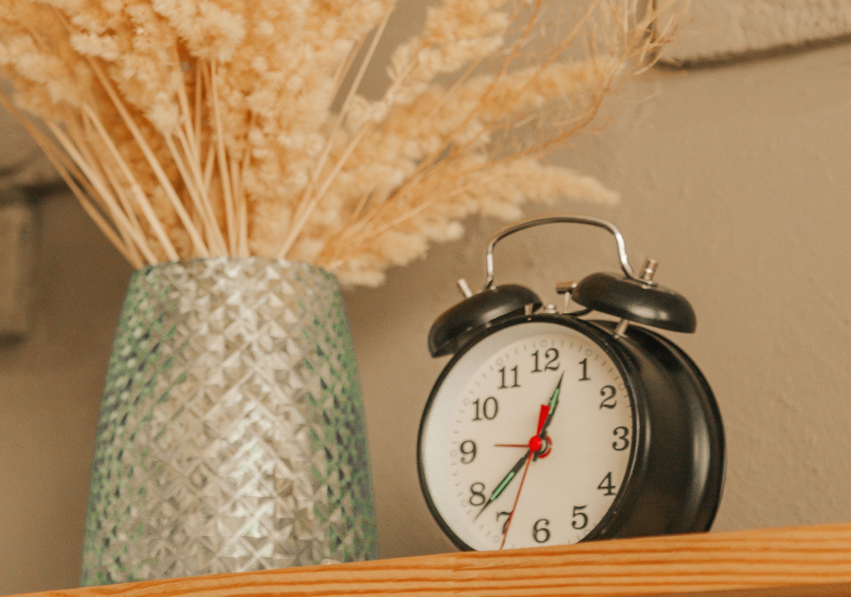 A bedroom shelf with an alarm clock and some dried plants in a vase.